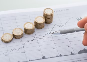 Cropped image of businessman's hand with pen analyzing line and coins graphs on desk